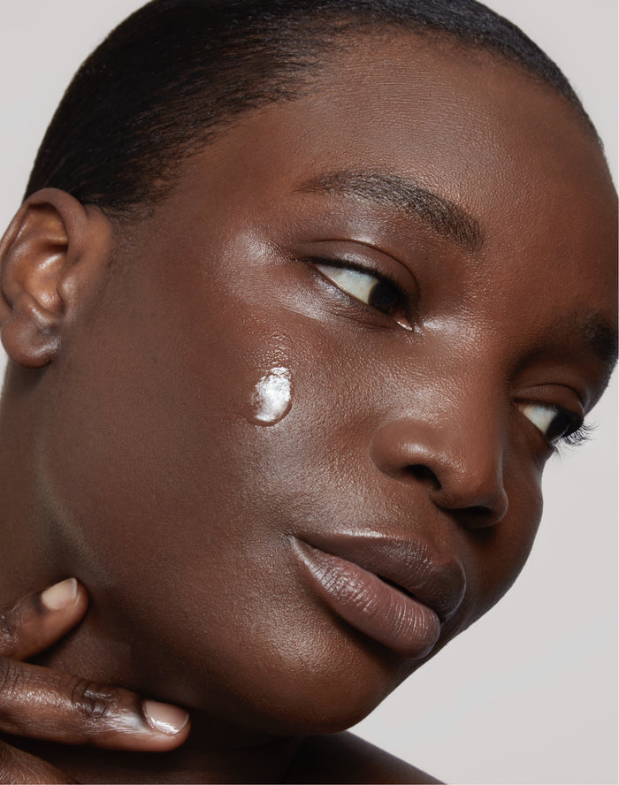 Una persona de piel lisa y oscura se aplica una gota de producto para el cuidado de la piel como parte de su rutina cuidado facial.