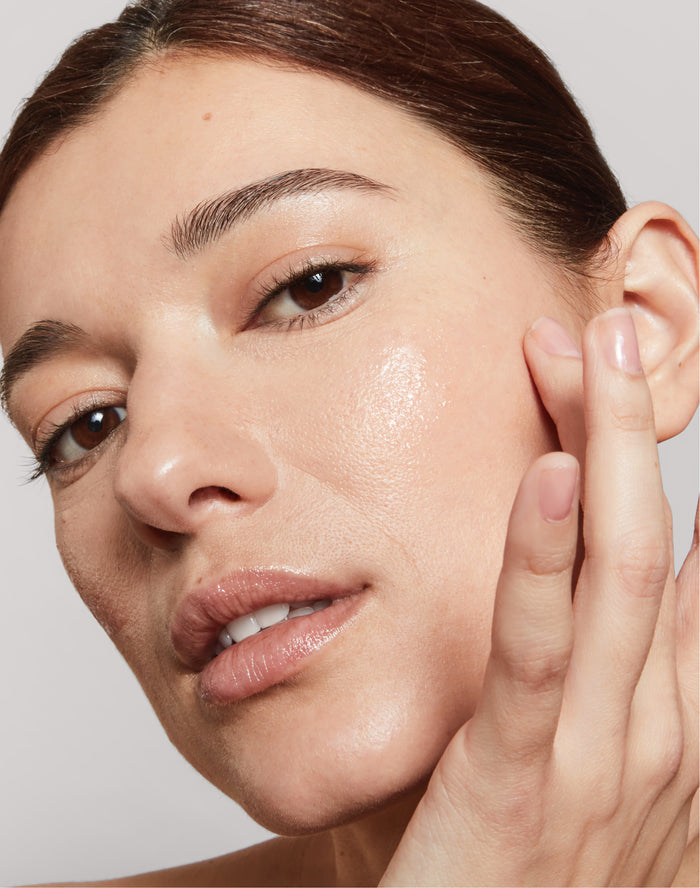 Una mujer se toca la mejilla, mostrando una piel hidratada gracias a un cuidado facial básico de rutina y un maquillaje neutro.
